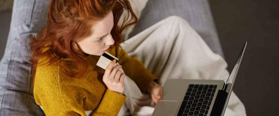 Young redhead woman relaxing on a sofa at home with her laptop computer on her lap as she shops online with her credit card reading information on the screen with an absorbed expression
