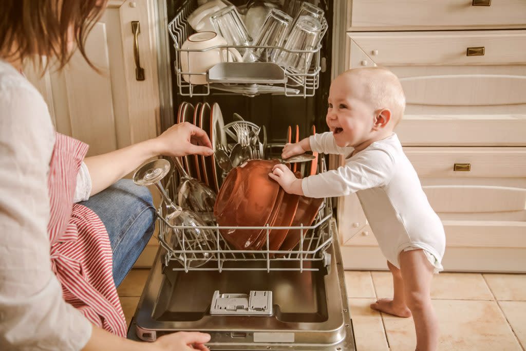 A baby leans on the dishwasher as their mother unloads it. 