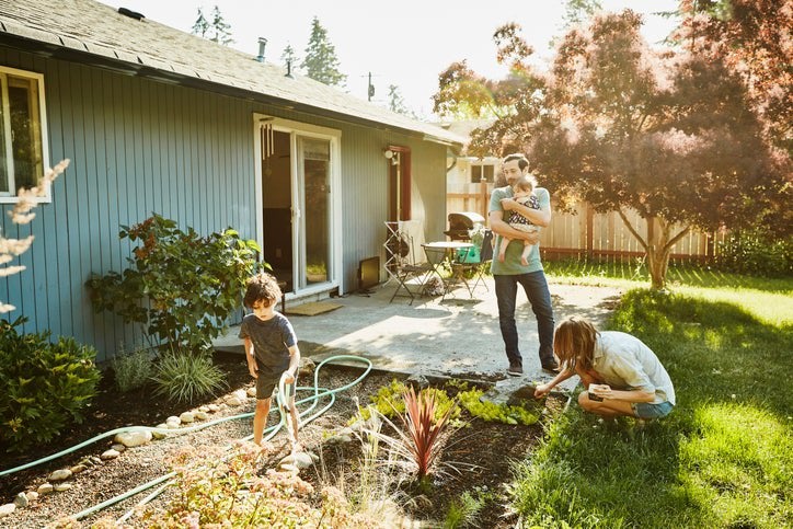 A mom working in the backyard garden with a toddler while the dad walks out of the house holding a baby.